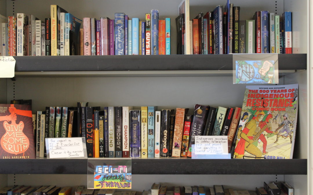 A landscape photo of a section of the middle school library. There is colorful, hand-drawn genre sign for the Sci-Fi Thriller section.